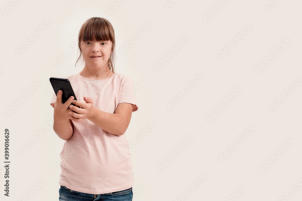 Portrait of disabled girl with Down syndrome looking at camera while using smartphone, standing isolated over white background Portrait of disabled girl with Down syndrome looking at camera while using smartphone, standing isolated over white background