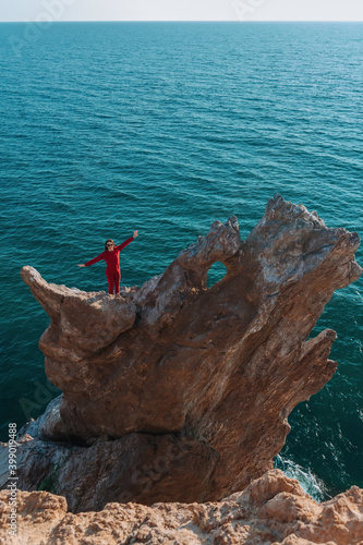 A beautiful girl with long dark hair and red clothes stands astride a huge rock in the shape of a dragon on the background of the sea. Her hands are up. The concept of active recreation.