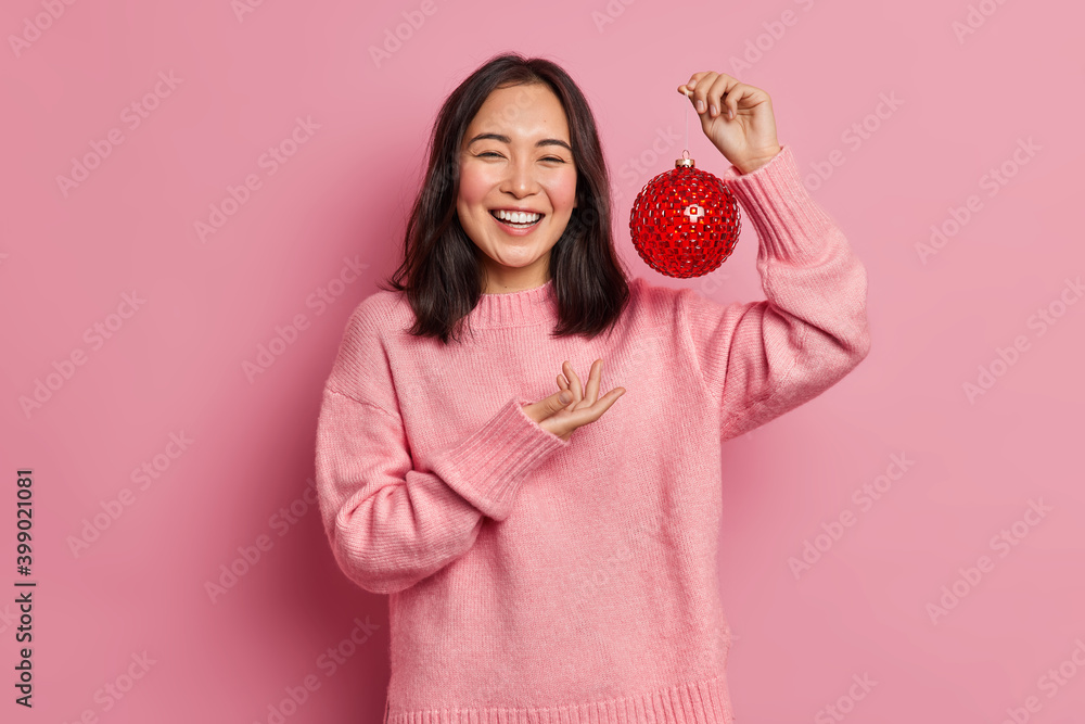 Smiling positive Asian woman with dark hair holds glass bauble for decorating Christmas tree enjoys buying new year attributes awaits for holidays wears casual jumper poses indoor over pink wall Smiling positive Asian woman with dark hair holds glass bauble for decorating Christmas tree enjoys buying new year attributes awaits for holidays wears casual jumper poses indoor over pink wall