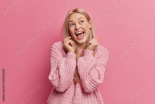 Overjoyed blonde young woman has happy face expression looks above keeps mouth opened dressed in casual clothes isolated over pink background. Attractive European girl imagines something funny