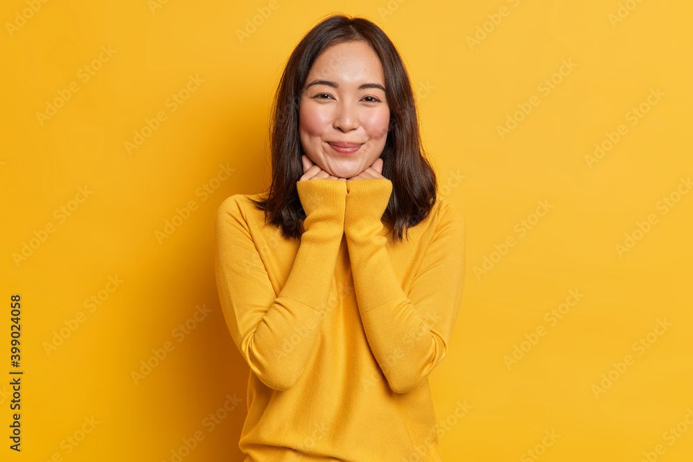 Studio shot of pretty young Asian woman keeps hands under chin has satisfied expression wears casual clothes isolated over yellow vivid background. Monochrome shot. Smiling eastern female model Studio shot of pretty young Asian woman keeps hands under chin has satisfied expression wears casual clothes isolated over yellow vivid background. Monochrome shot. Smiling eastern female model