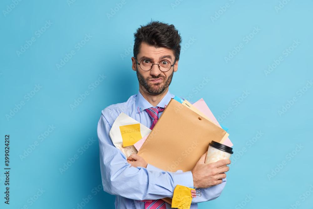 Displeased overworked student works hard before examination session holds disposable cup of coffee and papers remembers about deadline wears formal clothes isolated over blue studio background Displeased overworked student works hard before examination session holds disposable cup of coffee and papers remembers about deadline wears formal clothes isolated over blue studio background