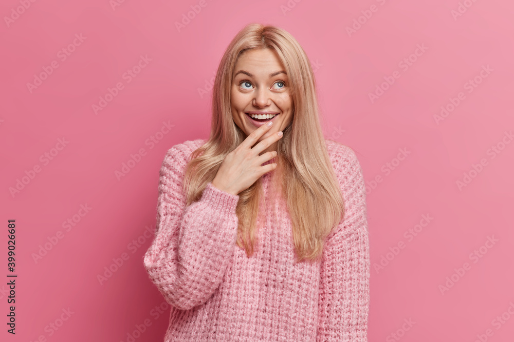Joyful blonde woman with thoughtful happy expression concentrated above smiles broadly shows white teeth wears warm winter sweater isolated over pink background. Positive human emotions concept. Joyful blonde woman with thoughtful happy expression concentrated above smiles broadly shows white teeth wears warm winter sweater isolated over pink background. Positive human emotions concept.