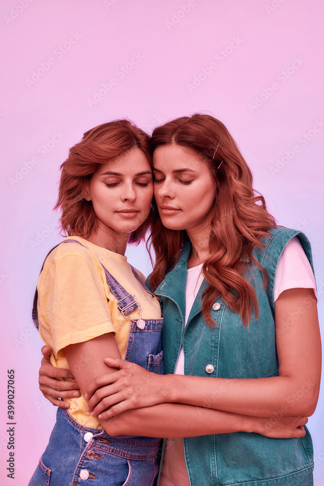Portrait of two attractive young girls, twin sisters holding each other, posing together with eyes closed isolated over pink background Portrait of two attractive young girls, twin sisters holding each other, posing together with eyes closed isolated over pink background
