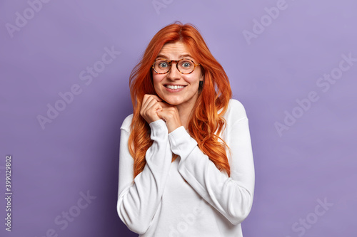 Portrait of cheerful surprised redhead European woman keeps hands together smiles toothily wears transparent glasses and white jumper reacts on pleasant revelation isolated over purple background