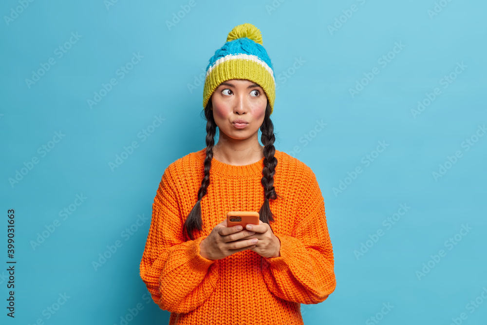 Φωτογραφία Photo of lovely brunette woman has two pigtails purses lips holds mobile phone sends text messages enjoys online communication dressed in warm knitted jumper and hat poses indoor