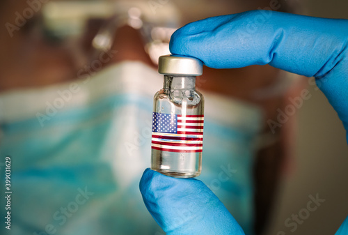 Close-up of medical vial with the flag of United States and a blurred doctor on the background. Selective focus. Concept of Covid vaccination campaign in the USA
