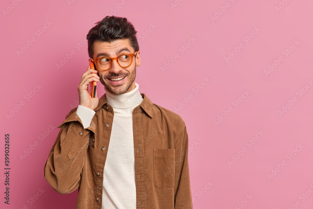Unshaven positive European adult man has telephone conversation keeps cellular near ear enjoys pleasant talk with colleague wears optical glasses brown shirt poses indoor with blank space aside Unshaven positive European adult man has telephone conversation keeps cellular near ear enjoys pleasant talk with colleague wears optical glasses brown shirt poses indoor with blank space aside