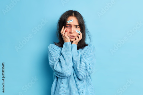 Depressed woman with bruise becomes victim of domestic rape violence suffers sexual abuse keeps hands on face wears knitted sweater isolated overblue background. Asian female being hit by cruel person