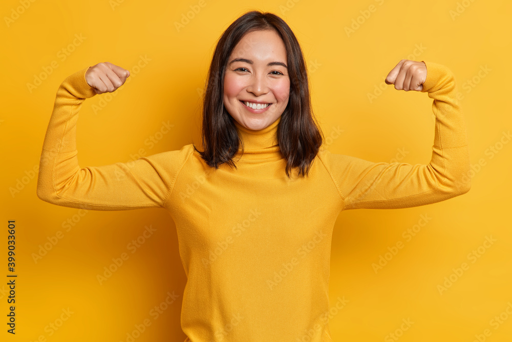 Pretty young brunette Asain woman raises arms and shows biceps demonstrates muscles feels proud about personal achievements wears yellow turtleneck stands indoor. People strength and power concept Pretty young brunette Asain woman raises arms and shows biceps demonstrates muscles feels proud about personal achievements wears yellow turtleneck stands indoor. People strength and power concept