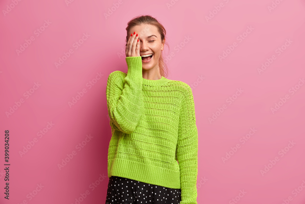 Φωτογραφία Studio shot of happy millennial girl makes face palm and laughs joyfully wears knitted jumper isolated over pink background