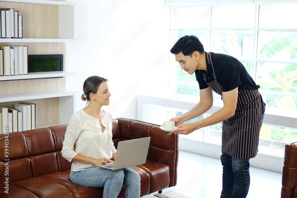 Smiling Asian barista young man or Waiter is Serving a cup of hot coffee for caucasian businesswoman is sitting on sofa and working online with laptop at modern cafe. Lifestyle and business Concept. Smiling Asian barista young man or Waiter is Serving a cup of hot coffee for caucasian businesswoman is sitting on sofa and working online with laptop at modern cafe. Lifestyle and business Concept.