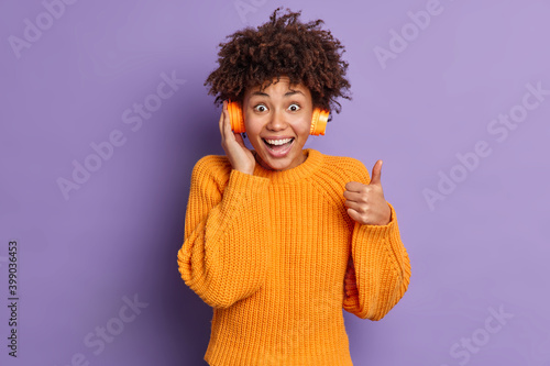 Positive millennial girl with curly Afro hair makes thumb up gesture rates song while listens audio track says thats excellent music wears knitted orange jumper isolated over purple background