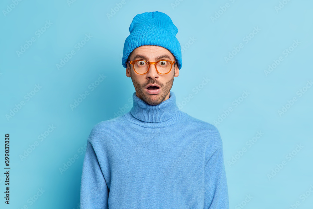 Portrait of surprised hipster guy keeps mouth opened from wonder wears hat and turtleneck isolated over blue background astonished over big news stares amazed. Omg no way. Human emotions concept