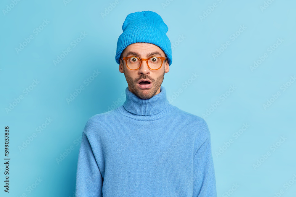 Portrait of surprised hipster guy keeps mouth opened from wonder wears hat and turtleneck isolated over blue background astonished over big news stares amazed. Omg no way. Human emotions concept Portrait of surprised hipster guy keeps mouth opened from wonder wears hat and turtleneck isolated over blue background astonished over big news stares amazed. Omg no way. Human emotions concept