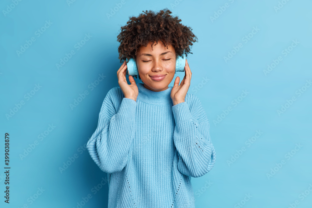 Φωτογραφία Studio shot of good looking woman with curly hair wears stereo electronics headphones enjoys listening music podcast wears warm sweater isolated over blue background