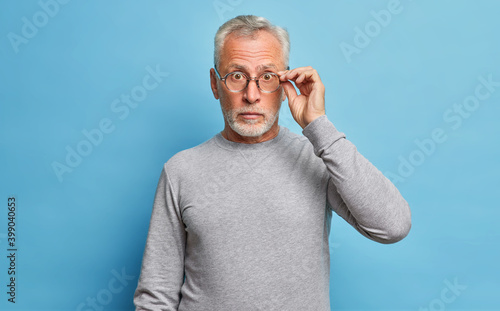 Surprised senior bearded Caucasian man stares through eyeglasses expresses shock wonders seasonal discounts and prices hears incredible news wears casual grey jumper isolated over blue background photo