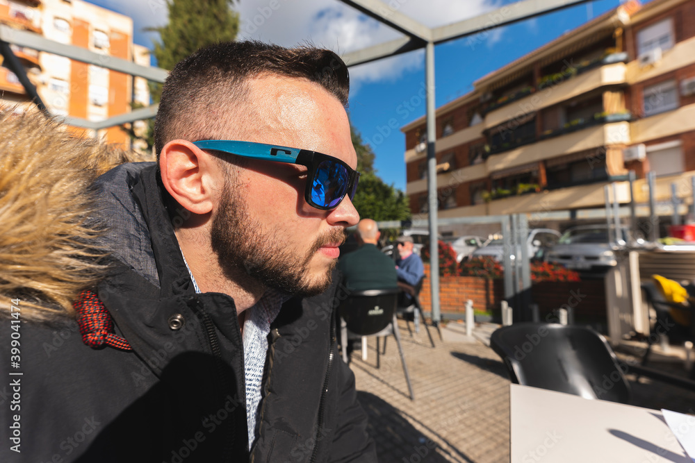 Scene on the bar terrace with a close-up of a young brown man of 25 years old wearing warm clothes and sitting in a Spanish tapas bar drinking beer and sunbathing with chairs in the background. Scene on the bar terrace with a close-up of a young brown man of 25 years old wearing warm clothes and sitting in a Spanish tapas bar drinking beer and sunbathing with chairs in the background.