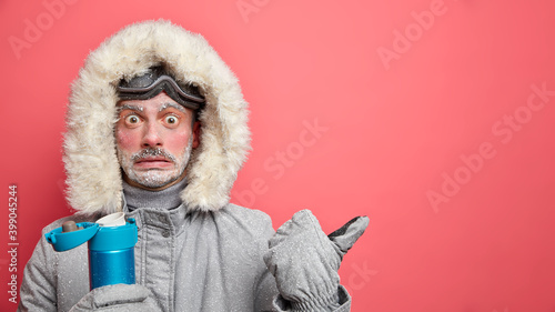 Wondered man spends time outdoors during blizzard and very cold day drinks hot beverage from thermos dressed in warm outerwear indicates aside on blank space attracts your attention to something photo