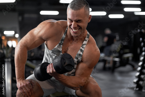 Muscular athletic bodybuilder fitness model sitting bench training biceps lift dumbbells in indoor gym