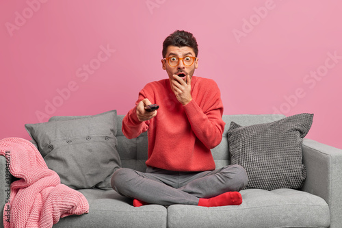 Photography Impressed stunned adult man stares at camera watches horrible movie holds remote control sits in lotus pose on comfortable sofa at living room against pink background