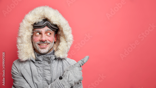 People recreation and winter rest concept. Positive frozen male skier dressed in warm thermo jacket and gloves points away on blank copy space and shows direction to cool seasonal resort place