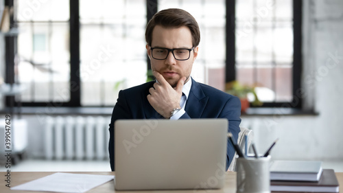 Close up thoughtful businessman wearing suit and glasses pondering project strategy, pensive young entrepreneur looking at laptop screen, touching chin, working on online project, financial stats