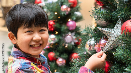 One happy Asian boy is enjoy Christmas decoration with many colorful ornaments on the pine tree. Kid and New year happiness concept. Kid decorates the star and balls with fun and joy.
