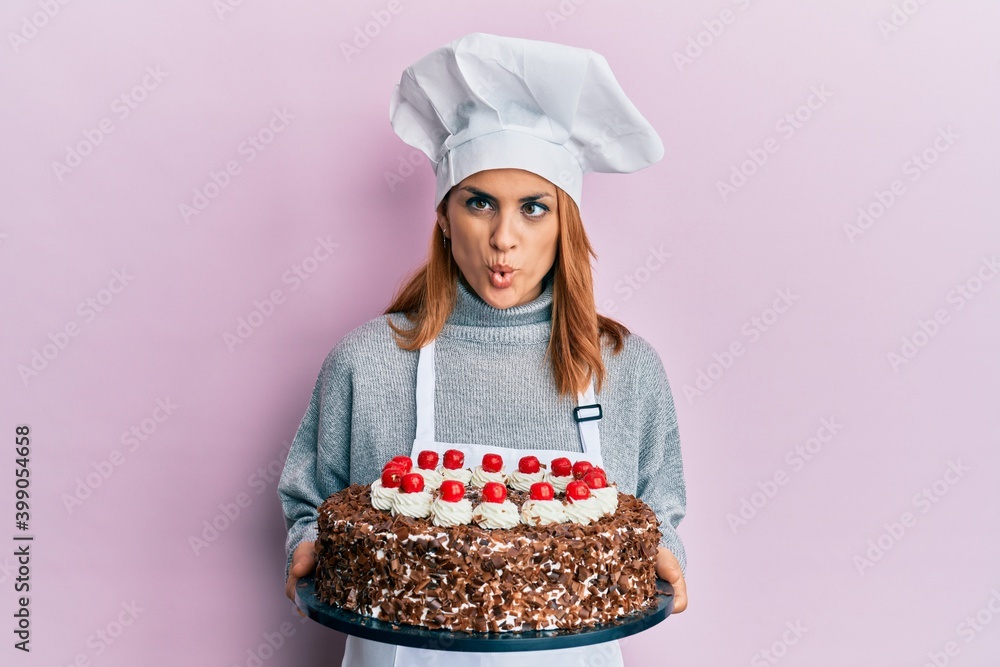 Hispanic young chef woman holding chocolate cake making fish face with mouth and squinting eyes, crazy and comical. Hispanic young chef woman holding chocolate cake making fish face with mouth and squinting eyes, crazy and comical.
