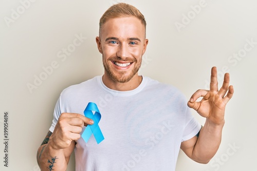 Young caucasian man holding blue ribbon doing ok sign with fingers, smiling friendly gesturing excellent symbol