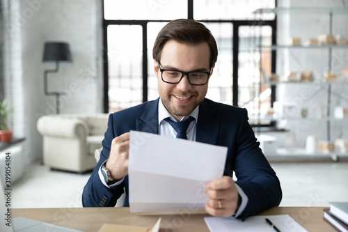 Close up overjoyed businessman reading good news in letter, holding paper sheet, sitting at desk in modern office, excited employee received job promotion, money refund, business achievement
