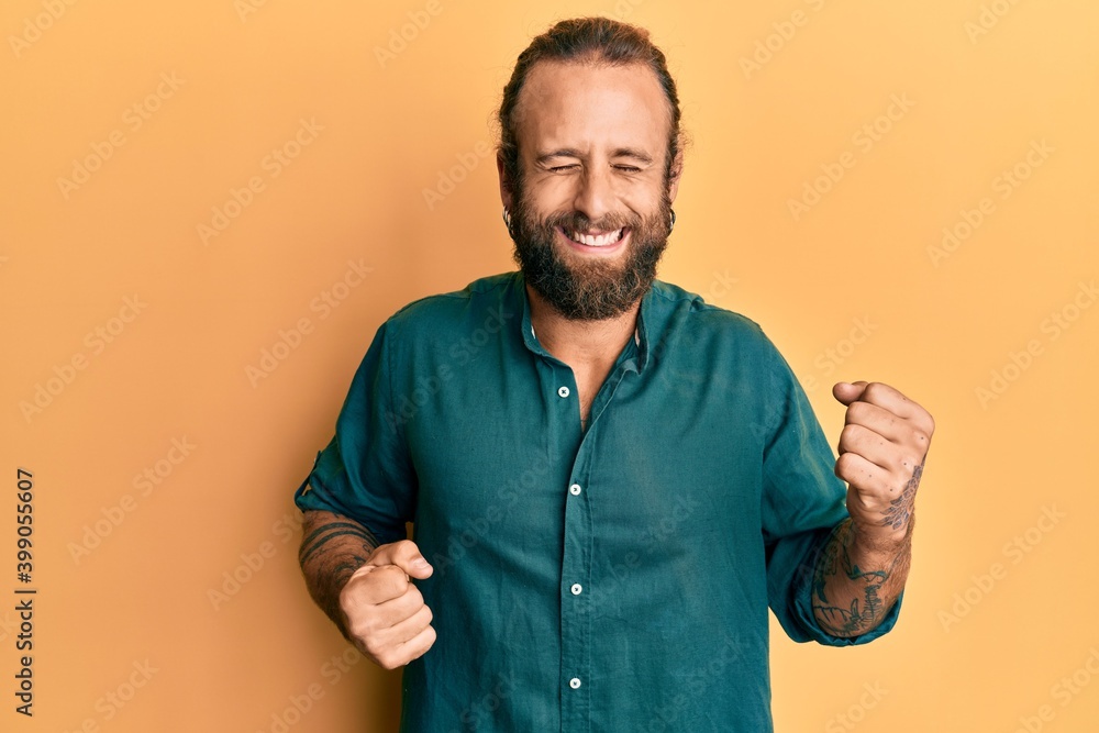 Handsome man with beard and long hair wearing casual clothes celebrating surprised and amazed for success with arms raised and eyes closed Handsome man with beard and long hair wearing casual clothes celebrating surprised and amazed for success with arms raised and eyes closed