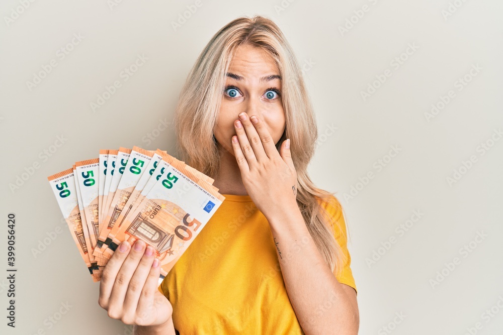Beautiful caucasian blonde girl holding bunch of 50 euro banknotes covering mouth with hand, shocked and afraid for mistake. surprised expression - obrazy, fototapety, plakaty 