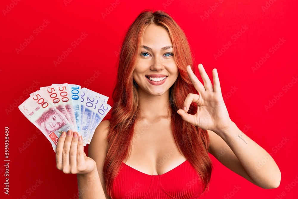 Young beautiful redhead woman holding swedish krona banknotes doing ok sign with fingers, smiling friendly gesturing excellent symbol Young beautiful redhead woman holding swedish krona banknotes doing ok sign with fingers, smiling friendly gesturing excellent symbol