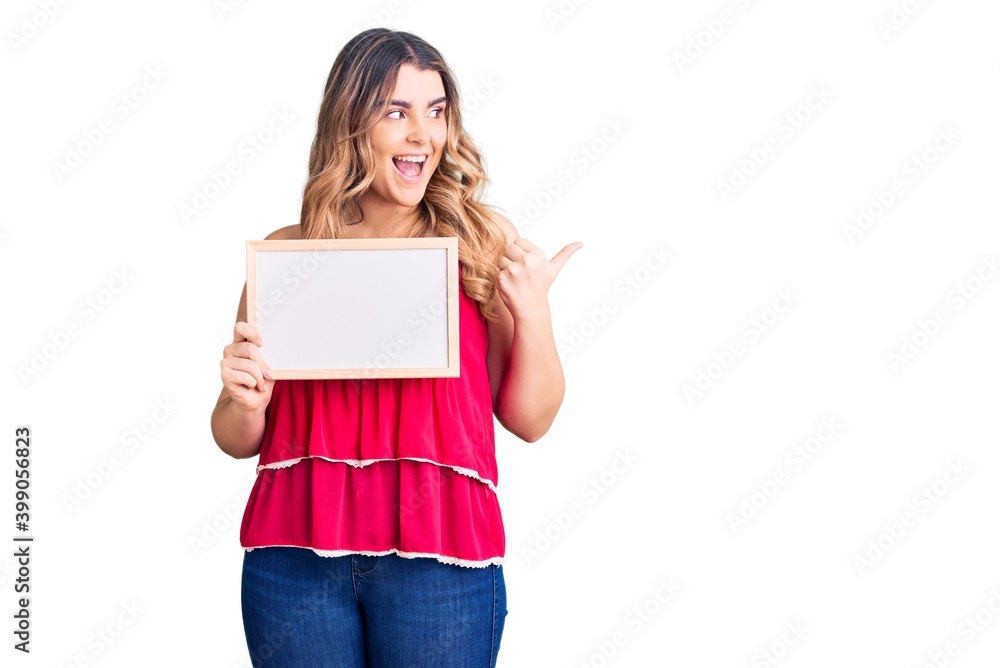 Young caucasian woman holding empty white chalkboard pointing thumb up to the side smiling happy with open mouth Young caucasian woman holding empty white chalkboard pointing thumb up to the side smiling happy with open mouth