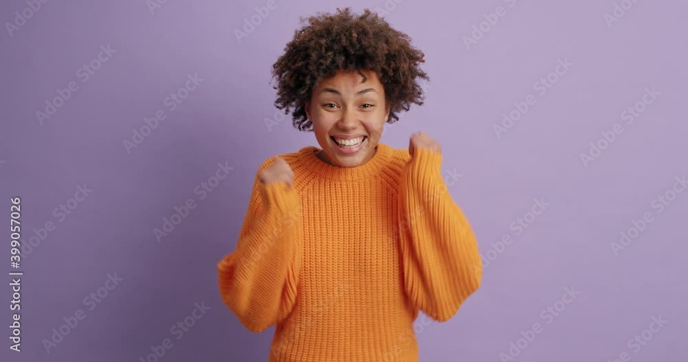 Happy dark skinned female model clenches fists celebrates awesome news wears orange knitted sweater smiles broadly feels like winner isolated over purple background. Positive feelings concept Happy dark skinned female model clenches fists celebrates awesome news wears orange knitted sweater smiles broadly feels like winner isolated over purple background. Positive feelings concept