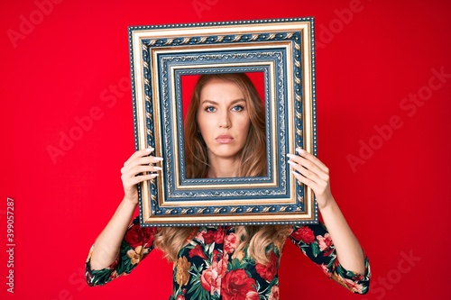 Young caucasian woman with blond hair holding empty frame relaxed with serious expression on face. simple and natural looking at the camera.