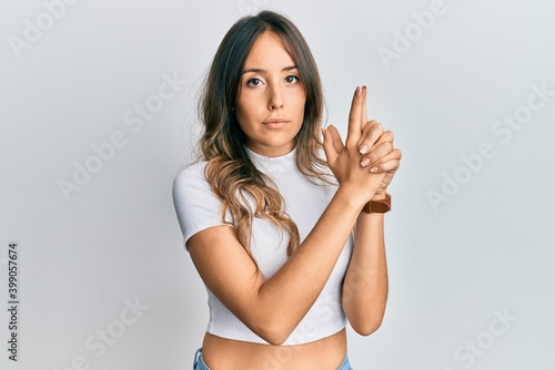 Young brunette woman wearing casual white t shirt holding symbolic gun with hand gesture, playing killing shooting weapons, angry face