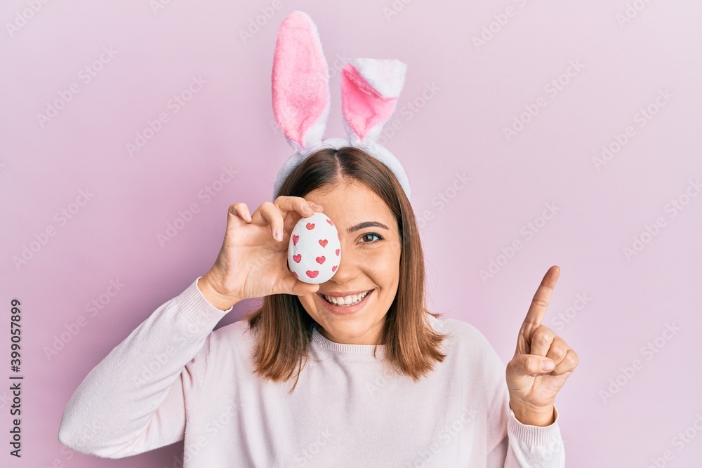 Young beautiful woman wearing cute easter bunny ears holding egg smiling with an idea or question pointing finger with happy face, number one Young beautiful woman wearing cute easter bunny ears holding egg smiling with an idea or question pointing finger with happy face, number one