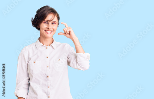 Beautiful young woman with short hair wearing elegant white shirt smiling and confident gesturing with hand doing small size sign with fingers looking and the camera. measure concept.