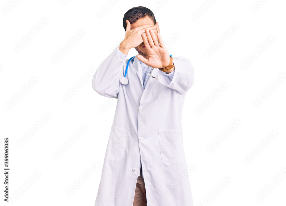 Handsome young man with bear wearing doctor uniform covering eyes with hands and doing stop gesture with sad and fear expression. embarrassed and negative concept. Handsome young man with bear wearing doctor uniform covering eyes with hands and doing stop gesture with sad and fear expression. embarrassed and negative concept.