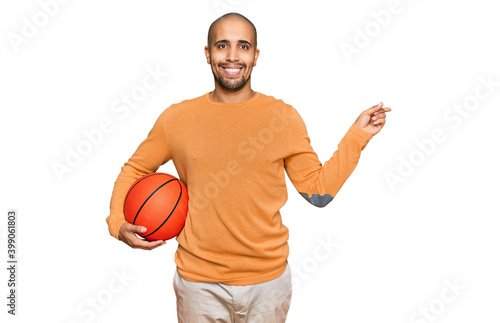 Hispanic adult man holding basketball ball smiling happy pointing with hand and finger to the side
