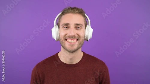 portrait of attractive laughing long-haired man wearing large white wireless headphones. on an isolated purple background. the man is wearing a burgundy sweater. 4K