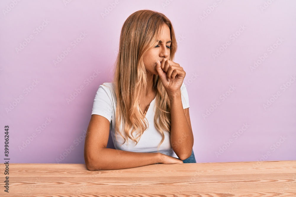Beautiful blonde young woman wearing casual clothes sitting on the table feeling unwell and coughing as symptom for cold or bronchitis. health care concept. Beautiful blonde young woman wearing casual clothes sitting on the table feeling unwell and coughing as symptom for cold or bronchitis. health care concept.