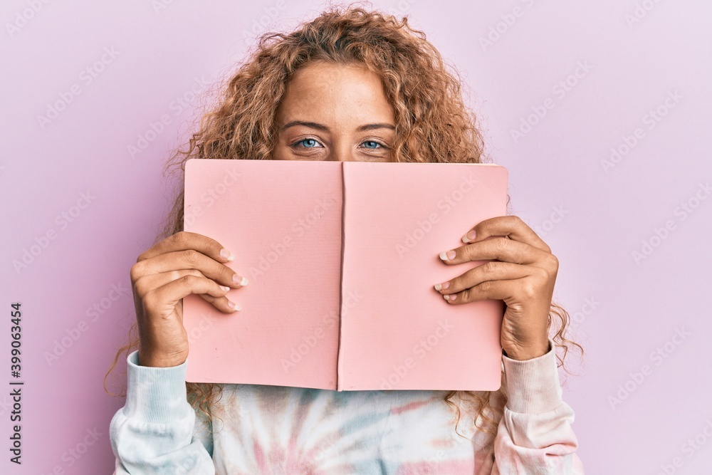 Beautiful caucasian teenager girl reading a book covering face smiling and laughing hard out loud because funny crazy joke. Beautiful caucasian teenager girl reading a book covering face smiling and laughing hard out loud because funny crazy joke.