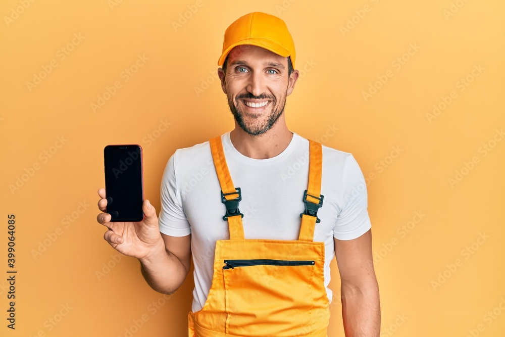 Young handsome man wearing handyman uniform showing smartphone screen looking positive and happy standing and smiling with a confident smile showing teeth Young handsome man wearing handyman uniform showing smartphone screen looking positive and happy standing and smiling with a confident smile showing teeth
