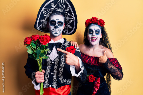 Young couple wearing mexican day of the dead costume holding roses smiling happy pointing with hand and finger