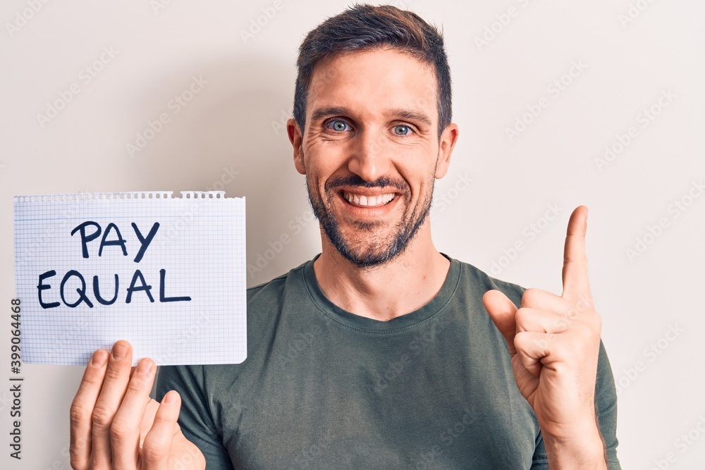 Young handsome man asking for equality economy holding paper with pay equal message smiling happy pointing with hand and finger to the side Young handsome man asking for equality economy holding paper with pay equal message smiling happy pointing with hand and finger to the side