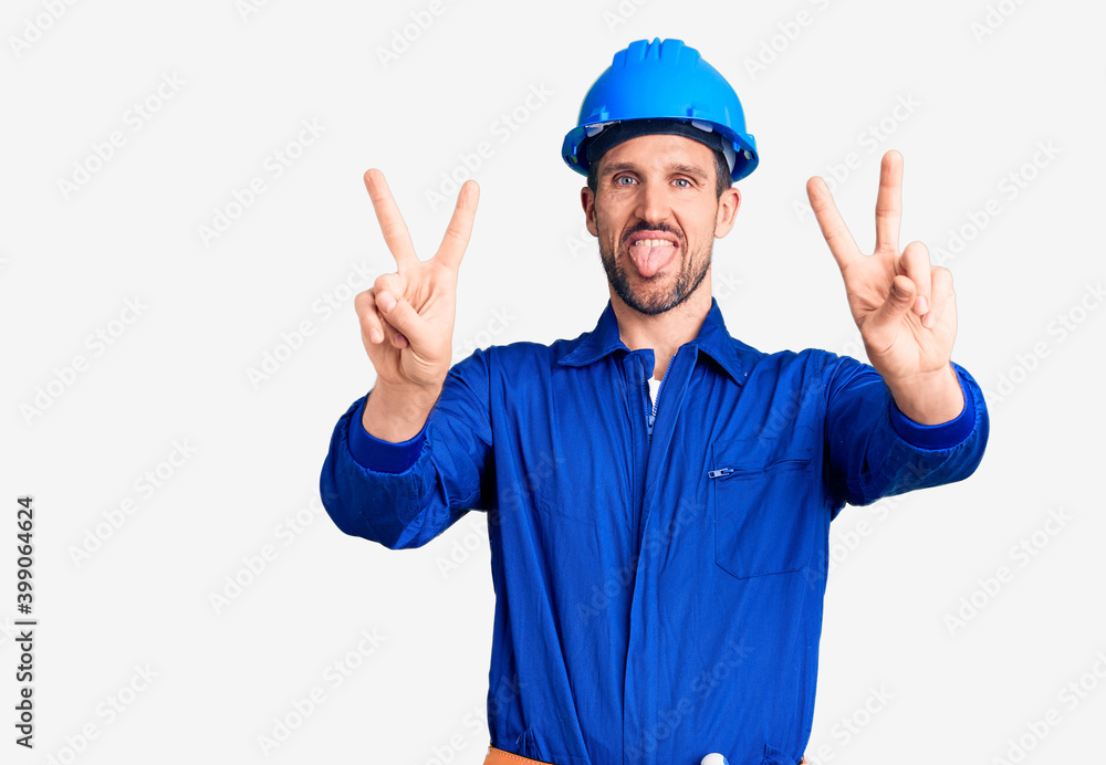 Young handsome man wearing worker uniform and hardhat smiling with tongue out showing fingers of both hands doing victory sign. number two.
