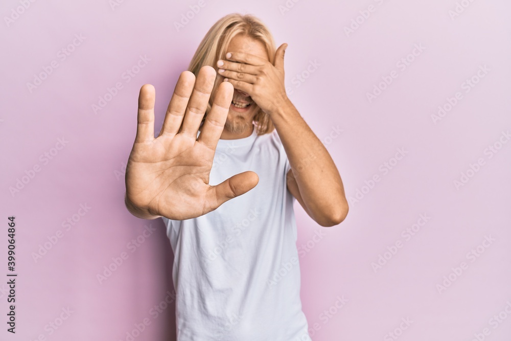 Caucasian young man with long hair wearing casual white t shirt covering eyes with hands and doing stop gesture with sad and fear expression. embarrassed and negative concept.